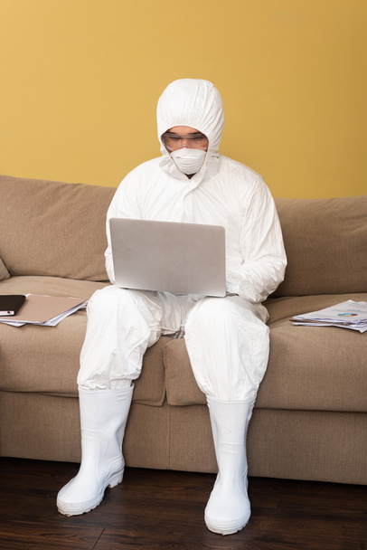 Mann in Schutzanzug, Schutzmaske und Schutzbrille mit Laptop in der Nähe von Dokumenten auf Couch  - Foto, Bild