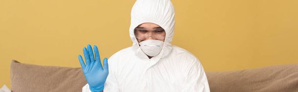 Orientação panorâmica do homem em terno hazmat, máscara médica e luva de látex acenando a mão em casa
  - Foto, Imagem