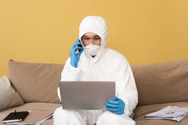 Freelancer em terno hazmat, luvas de látex e máscara médica falando no smartphone e usando laptop no sofá
  - Foto, Imagem