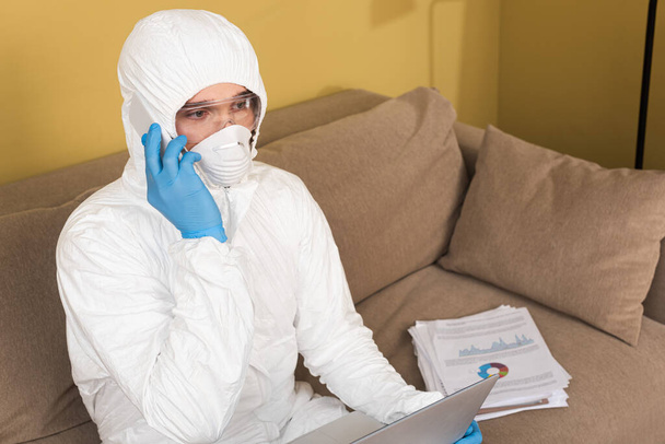 Freelancer in safety goggles, medical mask and hazmat suit talking on smartphone near laptop and documents on couch  - Foto, immagini