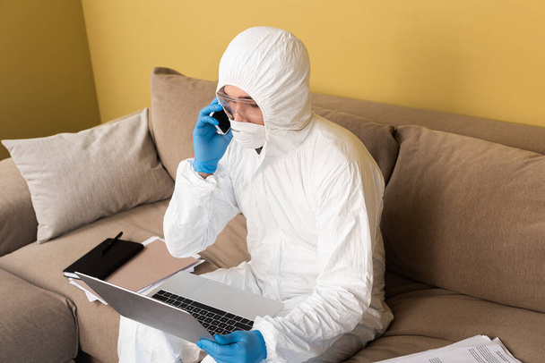Freelancer en traje de hazmat y guantes de látex hablando en smartphone y usando laptop en sofá en salón
 - Foto, Imagen