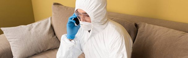 Panoramic crop of man in hazmat suit, medical mask and safety goggles talking on smartphone on couch  - Foto, Imagem