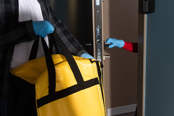 Przycięty widok dostawcę trzymającego torbę termo w pobliżu kobiety w rękawiczce lateksowej otwierającej drzwi - Zdjęcie, obraz
