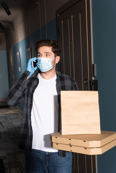 Entrega hombre en máscara médica sosteniendo cajas de pizza y paquete mientras habla en el teléfono inteligente en la entrada
 - Foto, imagen