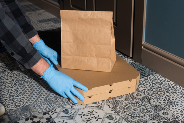 Обрезанный вид курьера в латексных перчатках кладет коробки пиццы и пакет на пол рядом с дверью
  - Фото, изображение