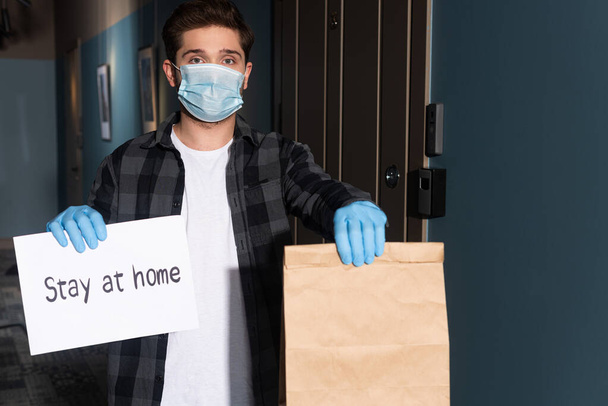Delivery man in medical mask and latex gloves holding package and card with stay at home lettering on entryway - Фото, изображение