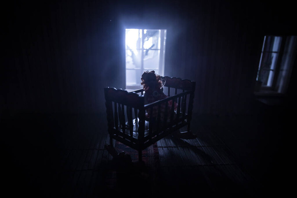 Παλιά ανατριχιαστική απόκοσμη κούνια μωρού κοντά στο παράθυρο σε σκοτεινό δωμάτιο. Τρομακτική σιλουέτα μωρού στο σκοτάδι. Ένα ρεαλιστικό σαλόνι κουκλόσπιτο με έπιπλα και παράθυρο τη νύχτα. Επιλεκτική εστίαση - Φωτογραφία, εικόνα