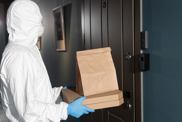 Oldalnézet futár vegyvédelmi ruha és orvosi maszk holding csomag és pizza dobozok ajtó közelében  - Fotó, kép