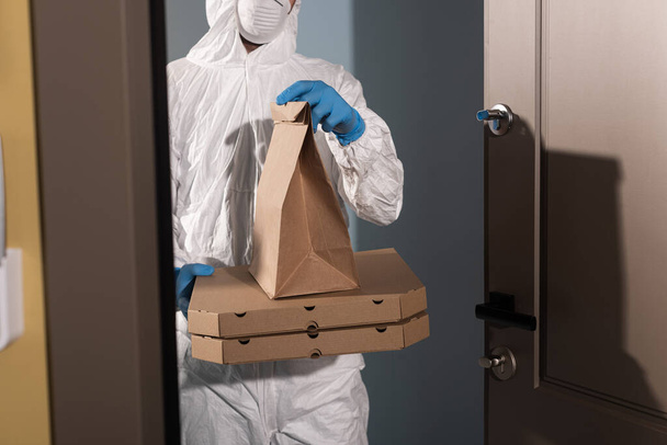 Vista recortada del repartidor con traje de materiales peligrosos y guantes de látex sosteniendo el paquete y cajas de pizza cerca de la puerta abierta
  - Foto, imagen