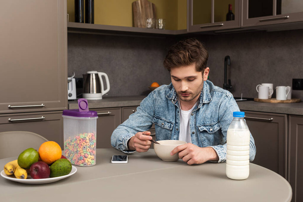 Homme mangeant des céréales près de fruits frais et smartphone sur la table dans la cuisine
 - Photo, image