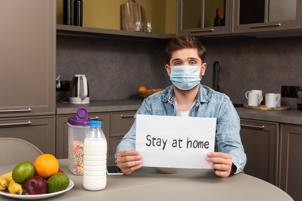 Man in medical mask holding card with stay at home lettering near cereals and fruits on table in kitchen  - Foto, Bild