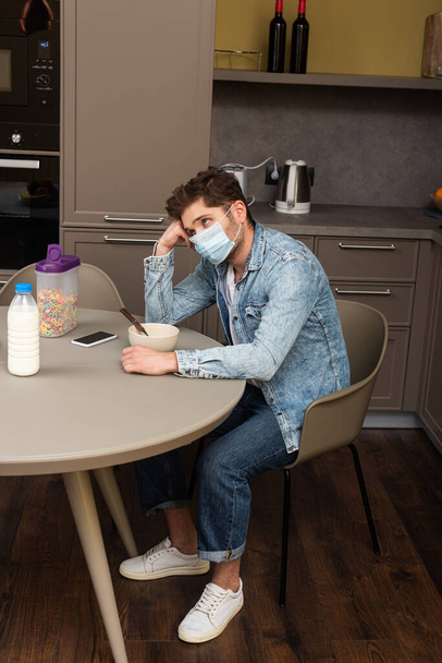 Tıbbi maskeli üzgün bir adamın mutfak masasındaki mısır gevreği ve akıllı telefonun yanından bakışları.  - Fotoğraf, Görsel