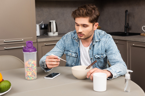 Hombre guapo sosteniendo termómetro cerca de teléfono inteligente y cereales en la mesa de la cocina
  - Foto, Imagen
