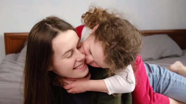 Cute adorable funny kid daughter kissing on cheek young mum playing on bed at home, smiling mother or nanny rest take, little girl enjoy tender moment with mom, love. Relative people family concept - Filmati, video