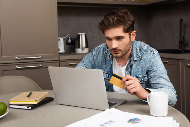 Selective focus of handsome man holding credit card and using laptop on kitchen table  - Foto, immagini