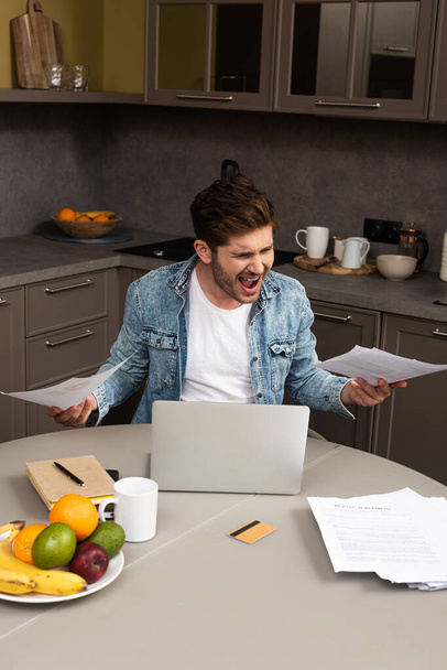 Mérges teleworker gazdaság dokumentumok közelében laptop és hitelkártya konyhaasztalon  - Fotó, kép