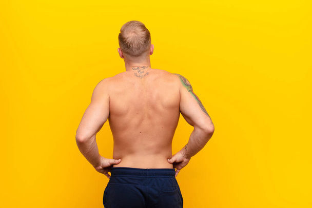 young strong blonde man feeling confused or full or doubts and questions, wondering, with hands on hips, rear view against yellow wall - Photo, image