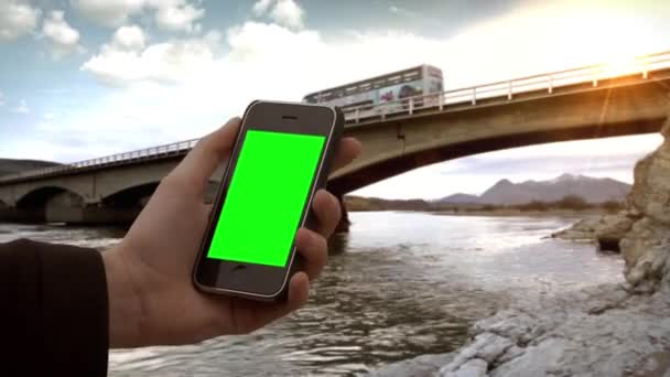 Mano masculina sosteniendo un Smartphone con pantalla verde bajo un puente en un río. Puede reemplazar la pantalla verde con las imágenes o imágenes que desee. Usted puede hacerlo con efecto de llave
. - Metraje, vídeo