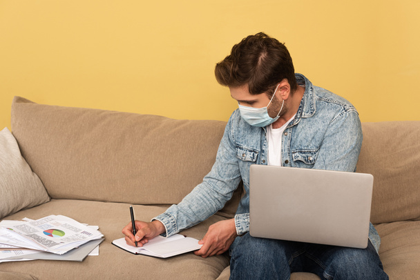 Фрилансер в медицинской маске пишет на ноутбуке рядом с ноутбуком и документы с картами на диване
  - Фото, изображение