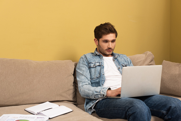 Jóképű szabadúszó használja laptop közelében papírok és notebook kanapén  - Fotó, kép
