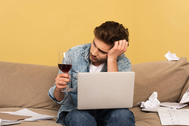 Töprengő szabadúszó kezében pohár bor közelében laptop és dokumentumok kanapén  - Fotó, kép