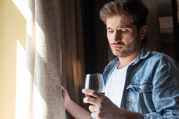 Zdenerwowany mężczyzna trzymający kieliszek wina przy oknie w salonie - Zdjęcie, obraz