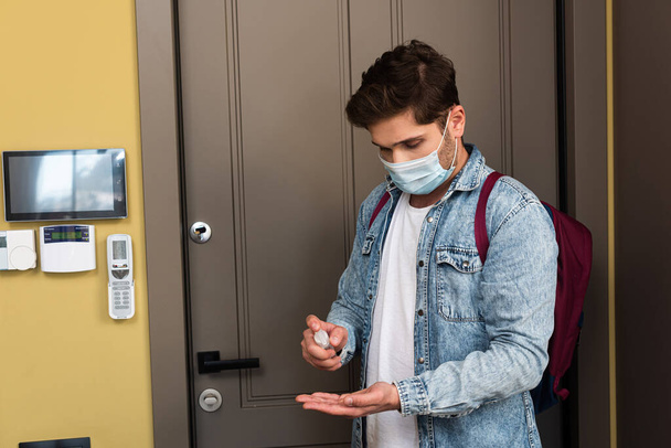 Junger Mann mit medizinischer Maske benutzt Händedesinfektionsmittel in der Nähe der Wohnungstür  - Foto, Bild