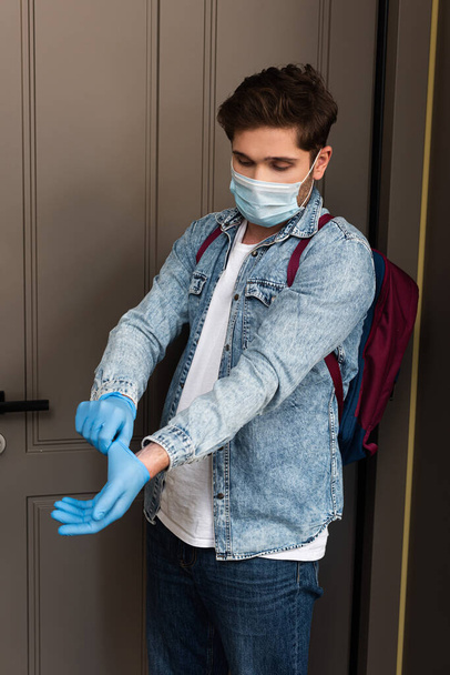Man in medical mask and backpack putting on latex gloves near door at home  - Fotografie, Obrázek