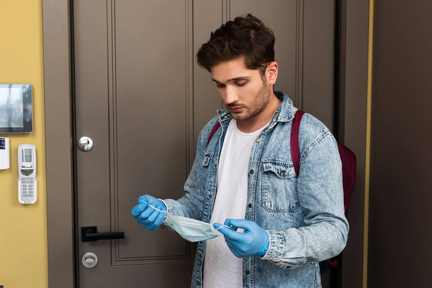 Handsome man in latex gloves holding medical mask near door in hallway  - 写真・画像