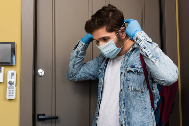 Young man in latex gloves putting on medical mask near door in hallway  - Fotoğraf, Görsel