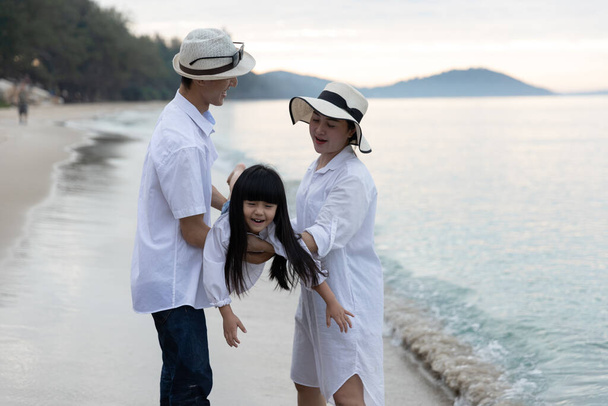 Ευτυχισμένη ασιατική οικογένεια στις καλοκαιρινές διακοπές Κόρη παίζει αεροπλάνο που φέρουν στην αγκαλιά πατέρες και η μητέρα μαζί στην παραλία το πρωί, την ανατολή του ηλίου. Holiday and Travel concept. - Φωτογραφία, εικόνα