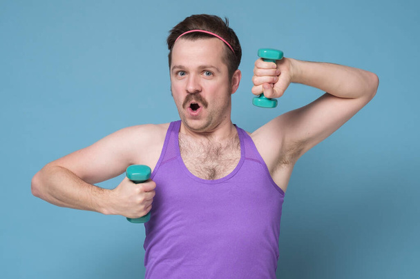 Satisfied young strength man with mustache lifting dumbbell isolated on blue background - Foto, Imagen