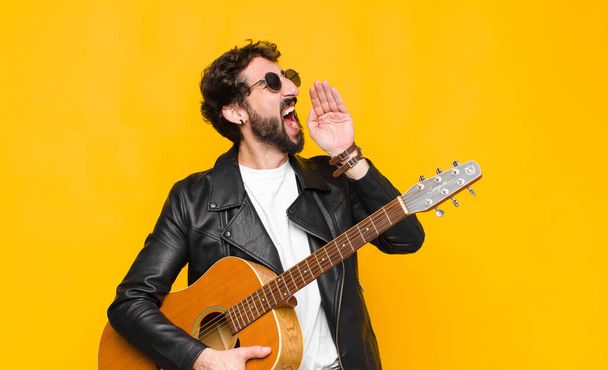 young musician man yelling loudly and angrily to copy space on the side, with hand next to mouth with a guitar, rock and roll concept - Фото, изображение