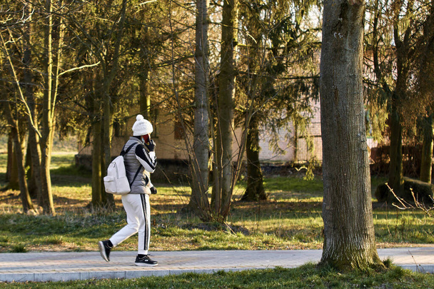 Ένα κορίτσι με άσπρη φόρμα περπατάει μόνο του στο πάρκο αναψυχής και μιλάει στο τηλέφωνο.. - Φωτογραφία, εικόνα