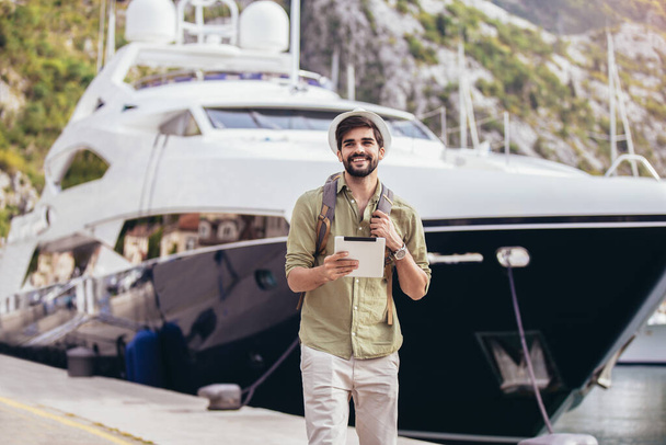 Young man walking by the harbor of a touristic sea resort with boats on background, holding digital tablet. - Foto, imagen