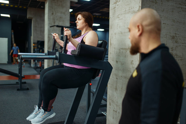 Κουρασμένη υπέρβαρο κορίτσι με εκπαιδευτή σε αθλητικό σύλλογο, προπόνηση φυσικής κατάστασης με εκπαιδευτή. Θηλυκό άτομο παλεύει με το υπερβολικό βάρος, αερόβια προπόνηση κατά της παχυσαρκίας, γυμναστήριο - Φωτογραφία, εικόνα