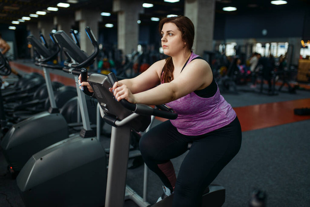 Femme en surpoids s'entraîne sur un vélo d'exercice dans la salle de gym, l'entraînement actif. Une femme obèse lutte contre l'excès de poids, l'entraînement aérobie contre l'obésité, club de sport
 - Photo, image