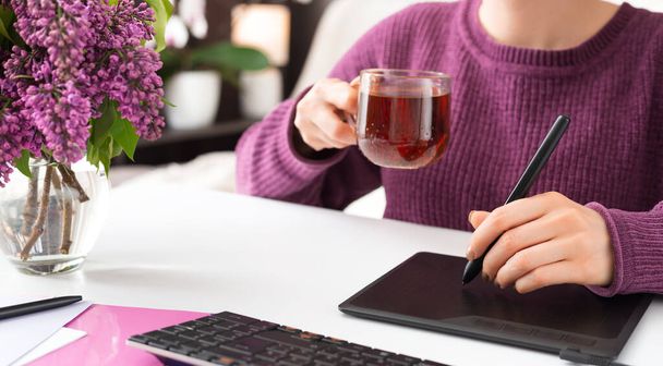 Graphic Designer retoucher freelancer works from home. Woman draws on graphic tablet in home office using laptop and computer. Remote work in cozy home office with flowers, cup of tea. - Foto, Imagem