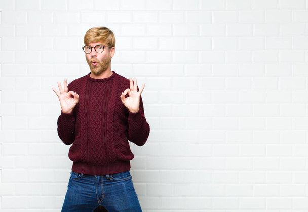 young blonde man feeling shocked, amazed and surprised, showing approval making okay sign with both hands agaist vintage tiles wall - Foto, Bild