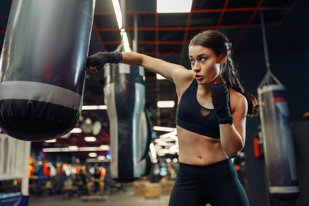 Woman in black boxing bandages and sportswear hits a punching bag, box training. Female boxer in gym, girl kickboxer in sport club, punches practice - Foto, afbeelding