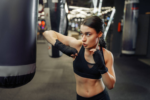Woman in black bandages and sportswear hits a punching bag, thai boxing training. Female boxer in gym, girl fighter in sport club, elbow punches practice - Zdjęcie, obraz
