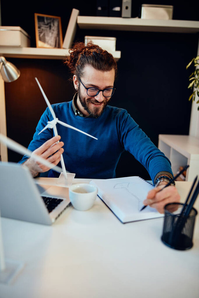 Mladý inovativní oddaný vousatý zaměstnanec sedí ve své moderní kanceláři a kreslí náčrtky větrných mlýnů. Koncept udržitelného rozvoje. - Fotografie, Obrázek