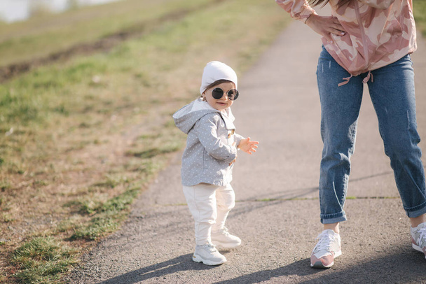 Feliz niña caminando con su madre en el parque. Mamá y su hija caminan cerca del paseo en el Día de las Madres. Linda niña en gafas de sol
 - Foto, imagen