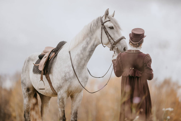 Девушка в старом костюме стоит рядом с серой лошадью в упряжке посреди высокой травы - вид сзади
 - Фото, изображение