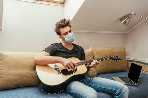young man in protective mask playing guitar while sitting on sofa near laptop with blank screen - Foto, Bild