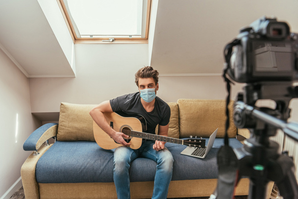 selectieve focus van jonge vlogger in medisch masker gitaar spelen terwijl het kijken naar digitale camera op statief - Foto, afbeelding