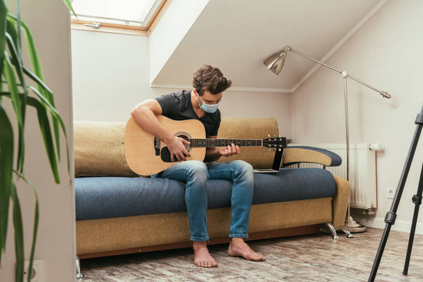 mladý muž v lékařské masce hraje na kytaru, zatímco sedí na pohovce v blízkosti notebooku - Fotografie, Obrázek