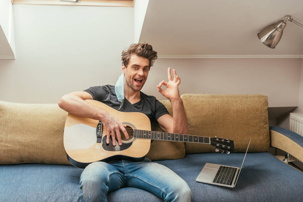 ενθουσιασμένος άνθρωπος με ιατρική μάσκα στο αυτί κρατώντας κιθάρα και δείχνει ροκ πινακίδα, ενώ κάθεται στον καναπέ κοντά στο laptop - Φωτογραφία, εικόνα