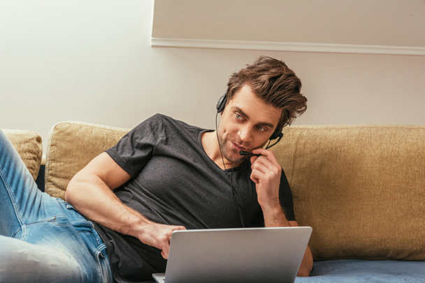 skeptical man in headset lying on sofa near laptop while working at home - Photo, Image