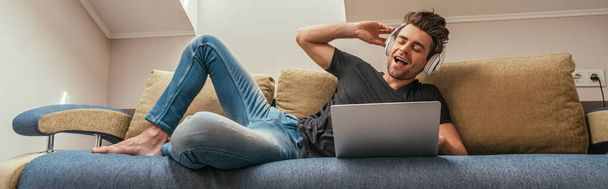 horizontal image of excited man listening music in wireless headphones while lying on sofa near laptop - Photo, Image